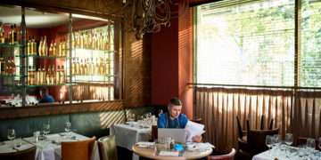 Mature man doing paperwork sitting at table with face mask and laptop, concentration, financial planning, organisation