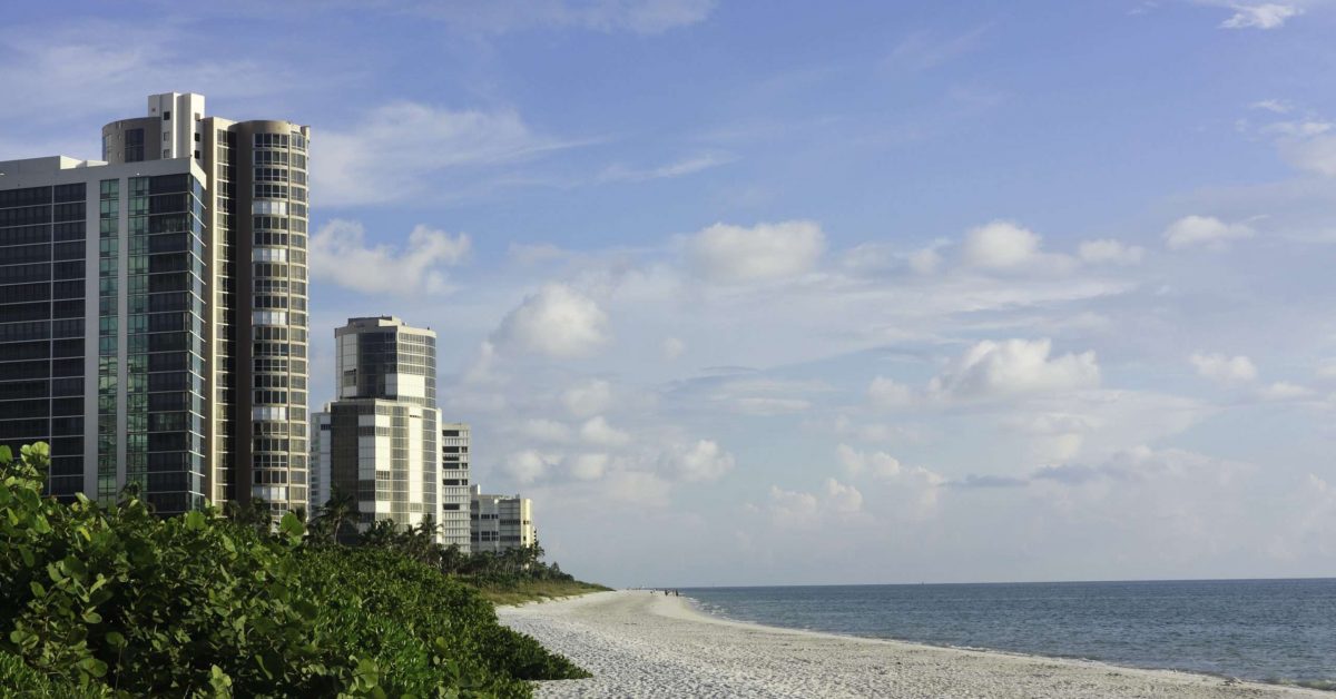 A wall of hotels and luxury condominiums lining the gulf coast of Florida at Naples Beach on the Gulf of Mexico.  Individuals in background not recognizable.